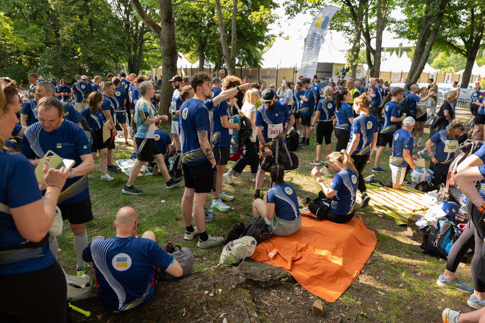 Eine Gruppe von DRV-Bund Mitarbeitenden in blauen DRV Bund T-Shirts versammelt sich im Freien. Einige sitzen auf einer orangefarbenen Decke oder auf dem Rasen, andere stehen. Im Hintergrund sind Bäume und Zelte zu sehen.