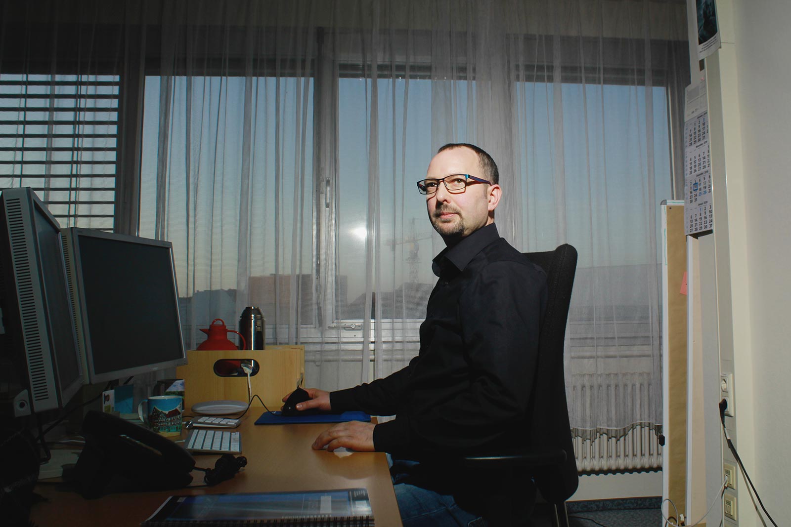 Eine männliche Person sitzt in einem Büro am Schreibtisch vor zwei Monitoren. Er trägt ein schwarzes Hemd und Brille. Die rechte Hand bedient die Maus.