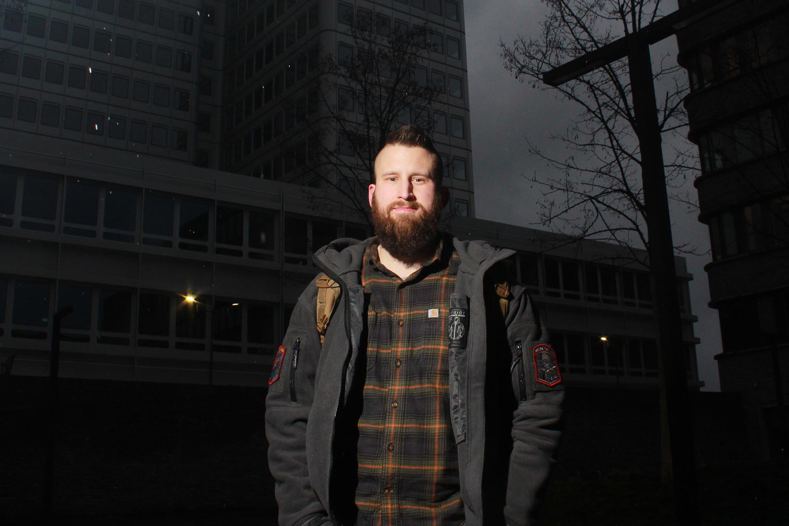 Eine männliche Person mit roten Haaren und Bart steht nachts vor dem Silberturm der DRV Bund in Berlin. Er trägt ein schwarz-orange kariertes Hemd und eine offene Winterjacke.