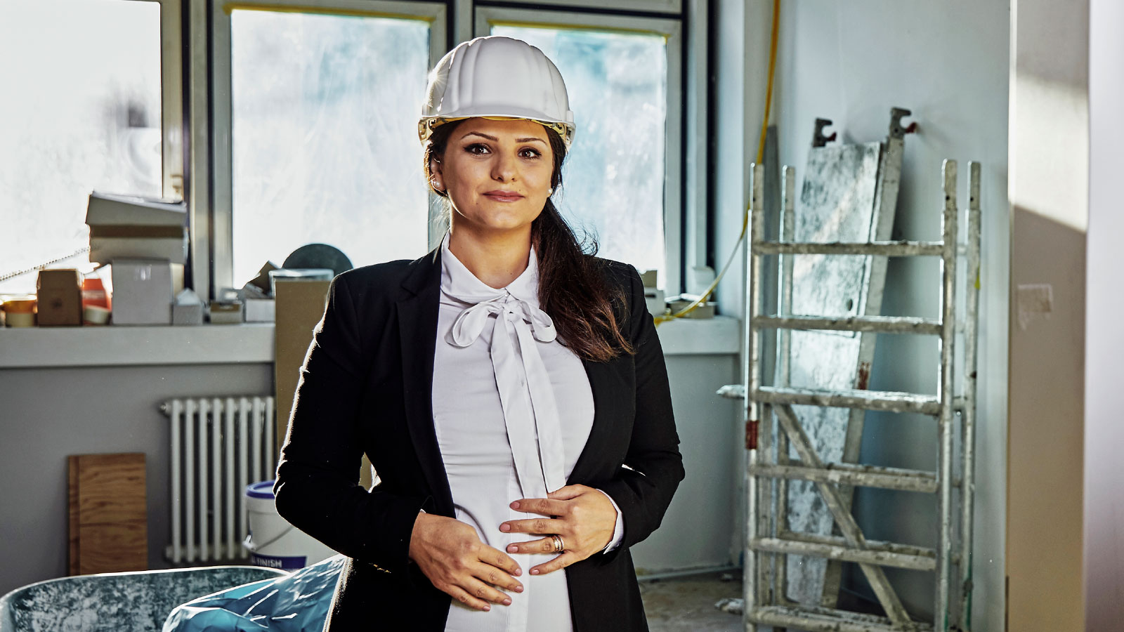 Eine weibliche Person steht in einem hellen Raum mit Baustellencharakter. Sie trägt weißen Schutzhelm, weißes Hemd mit Schleife und schwarzen Blazer. Die Hände sind vor dem Bauch verschränkt. Im Hintergrund sind Fenster, Leiter und Baumaterialien.