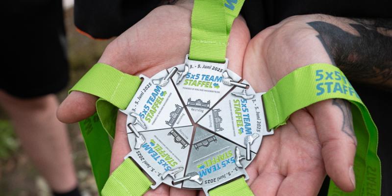 Nahaufnahme von Händen, die fünf zusammengesetzte Medaillen der 5x5 km Teamstaffel mit grünen Bändern halten. Die Medaillen ergeben zusammen einen Stern und tragen den Aufdruck „5x5 TEAM STAFFEL“.