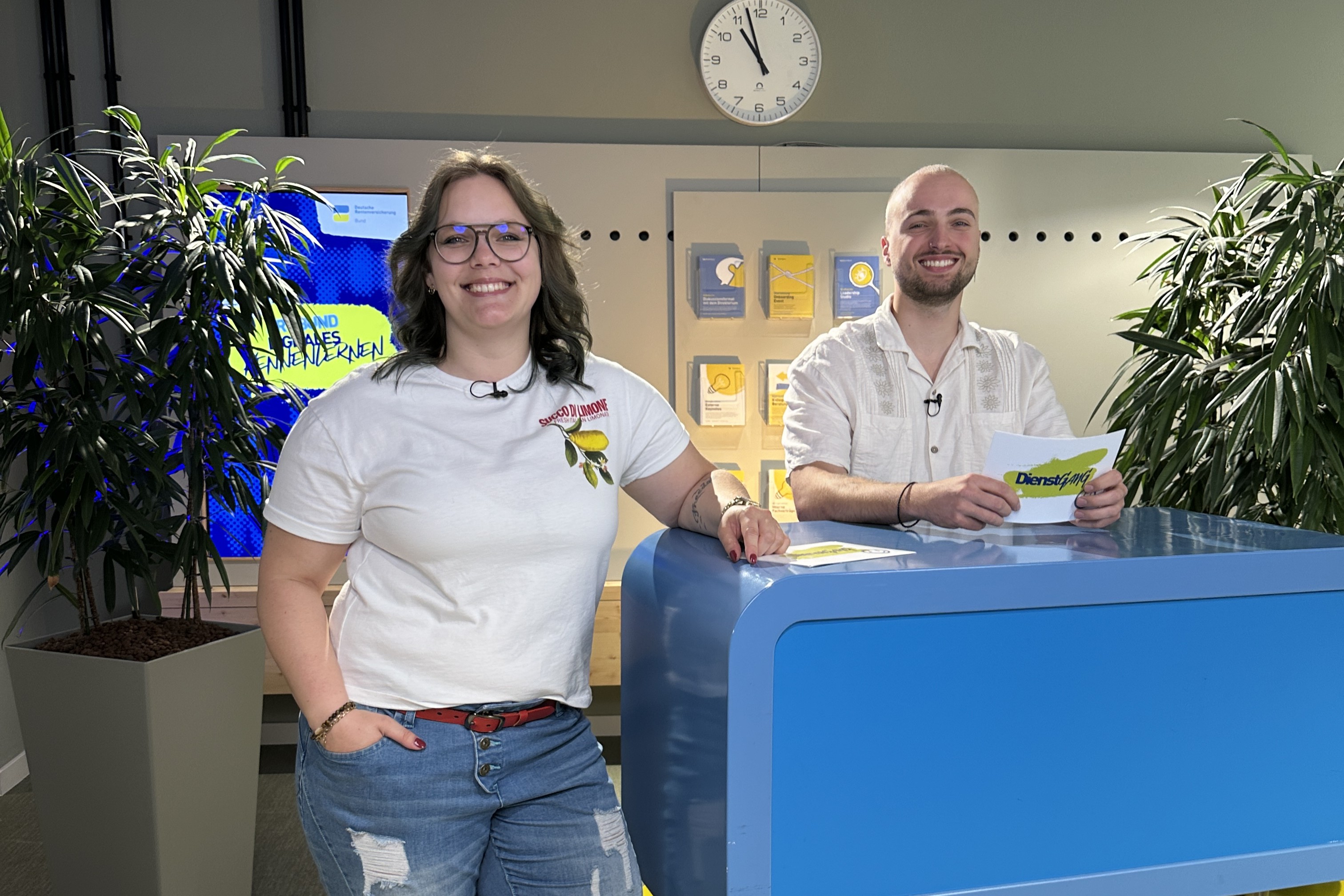 Zwei junge Menschen stehen lächelnd in einem modernen, freundlich gestalteten Studio. Die Frau steht , der Mann sitzt hinter einem blauen und gelben Moderationstisch mit Karten in der Hand. 