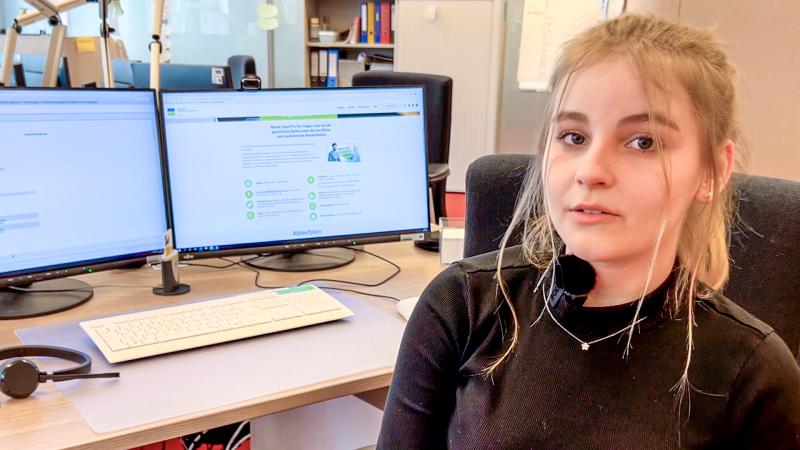 Auszubildende sitzt vor einem Schreibtisch im Büro, auf dem zwei Monitore mit Texten und Graphiken stehen.