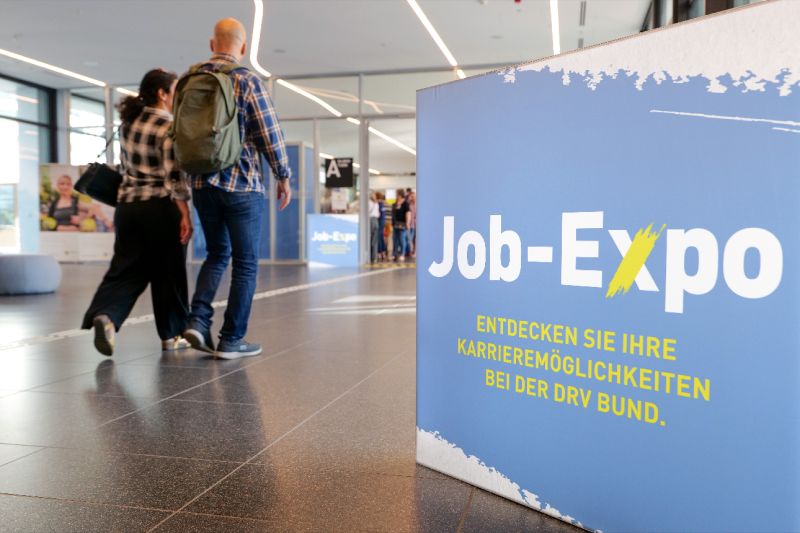 Zwei Personen bewegen sich durch eine Job-Expo. Im Vordergrund steht ein großes blaues Schild mit der Aufschrift: ‚Job-Expo – Entdecken Sie Ihre Karrieremöglichkeiten bei der DRV Bund.