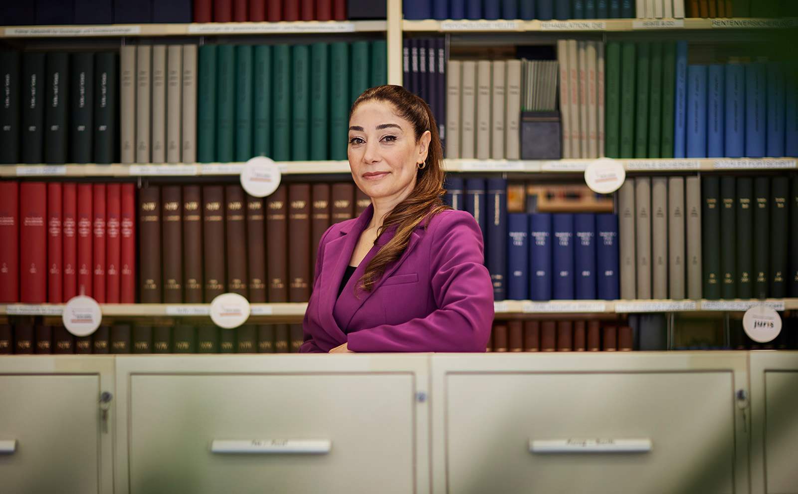 Frau Ören steht mit braunen, zum Zopf gebundenen Haaren in einem Büro. Sie trägt einen lilanen Anzug, stützt sich auf Aktenschränke und steht vor einer Bücherwand.