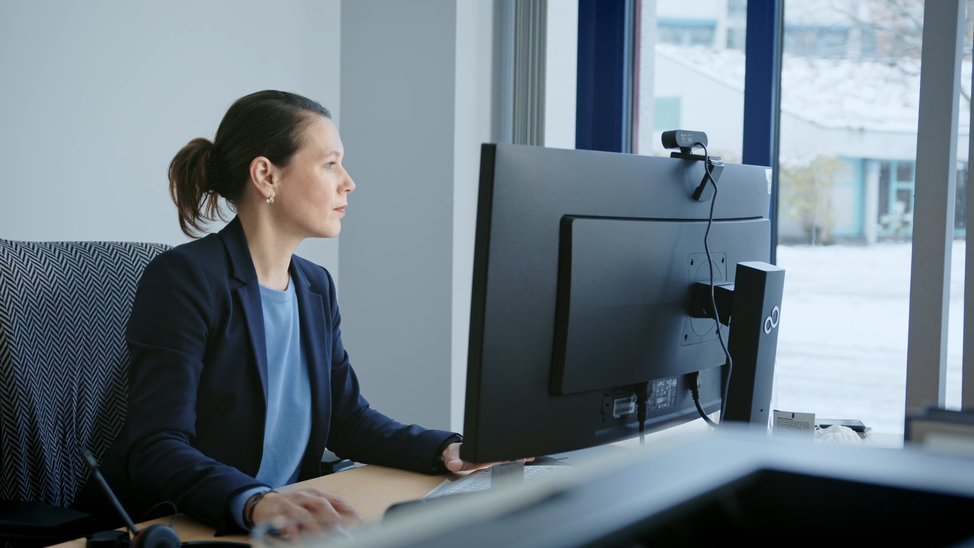 Eine Mitarbeiterin aus dem technischen Bereich sitzt in einem hellen Büro am Schreibtisch vor einem Computer und arbeitet konzentriert. Sie trägt ein blaues T-Shirt und einen dunkelblauen Blazer.