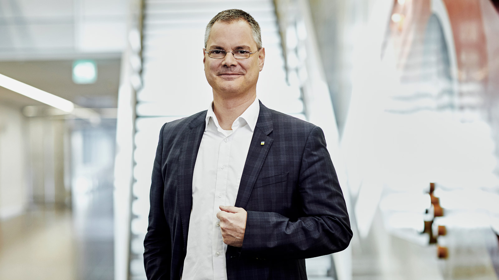 Dr. Jaster steht in einem dunklen Jackett und weißem Hemd vor einer weißleuchtenden Treppe in einem Bürogebäude.