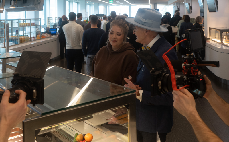 Eine DRV Mitarbeitende und Günther Krabbenhöft stehen in einer Kantine an; im Vordergrund zwei Personen und Kamerateam, links Obst- und Gemüseauslage.