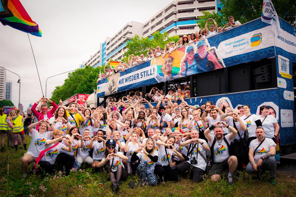Gruppe von Menschen mit Regenbogen-T-Shirts posiert vor bunt dekoriertem Bus mit Banner „NIE WIEDER STILL!“; im Hintergrund Wohngebäude und Bäume.